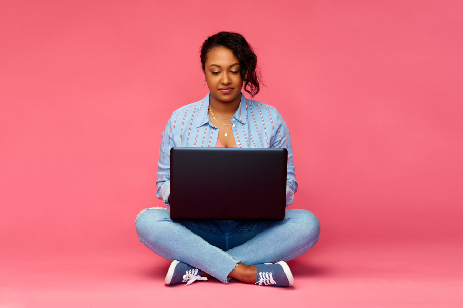 Young woman sits in front of a laptop on the floor, using AI. With a pink background.